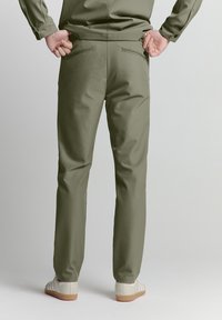 Olivgrüne Hose aus glattem Stoff, mit taillierter Passform, zwei Gesäßtaschen und kombiniert mit hellfarbenen Schuhen.
