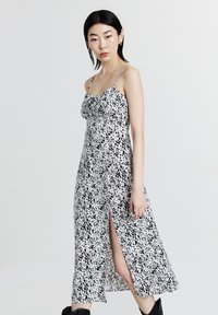 Donna che indossa un vestito midi senza maniche a pois bianchi e neri, con uno spacco laterale e stivali neri, su uno sfondo neutro.