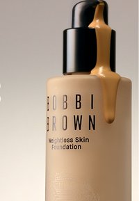 Bobbi Brown Weightless Skin Foundation i en klar glassflaske, med en svart pumpefugl og lys beige væske som dripper fra den.