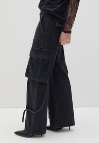 Pantalones cargo de pierna ancha en denim negro con múltiples bolsillos, una textura desgastada y un detalle de cordón en el dobladillo. Combinados con tacones de punta.
