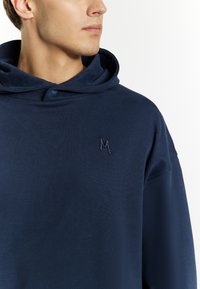 Mornarsko plava dukserica napravljena od mekanog materijala, s kapuljačom na vezivanje, dugmetom na izrezu i malim izvezenim logom "M" na prsima.