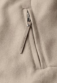 Beżowa tkanina o miękkiej fakturze z metalowym zamkiem i brązowym uchwytem zamka oznaczonym "StreetOne." Zbliżenie na szczegóły zamka.