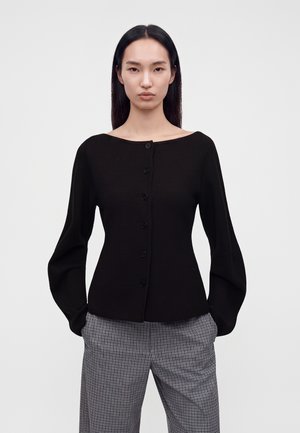 Vrouw met lang zwart haar, gekleed in een zwarte top met knopen en wijde mouwen en grijze geruite broek tegen een effen witte achtergrond.