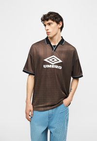 Mladi moški, ki nosi rjavo športno majico znamke Umbro z geometričnimi vzorci in svetlo modre obledene kavbojke, stoji z eno roko v žepu.