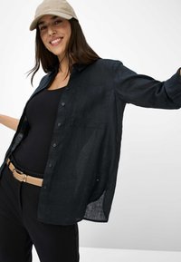 Schwarze Button-Down-Bluse mit lockerer Passform, aus leichtem Material. Hat eine Brusttasche und hochgerollte Ärmel. Wird über einem schwarzen Top getragen.