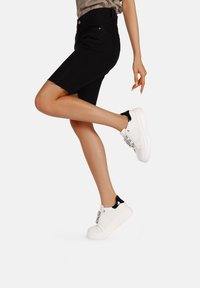 Sneaker bianchi con suola spessa, caratterizzati da un accentuato tacco nero e dettagli in catena. Indossati con una gonna di jeans nera a vita alta.