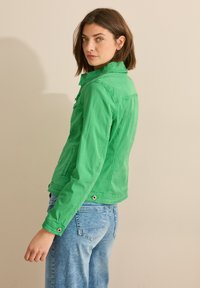 Femme avec les cheveux bruns courts portant une veste verte vif et un jean bleu clair, se tenant de côté et regardant par-dessus son épaule.