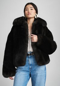 Μαύρο μπουφάν από ψεύτικη γούνα με φαρδύ γιακά, χαμηλωμένους ώμους και φουσκωτά μανίκια, φορεμένο πάνω από ένα ανοιχτόχρωμο τοπ και μπλε τζιν ψηλόμεσο.