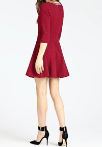 Robe rouge texturée à manches trois-quarts, jupe évasée et col rond. Style avec des chaussures noires à talons hauts munies de brides à la cheville.