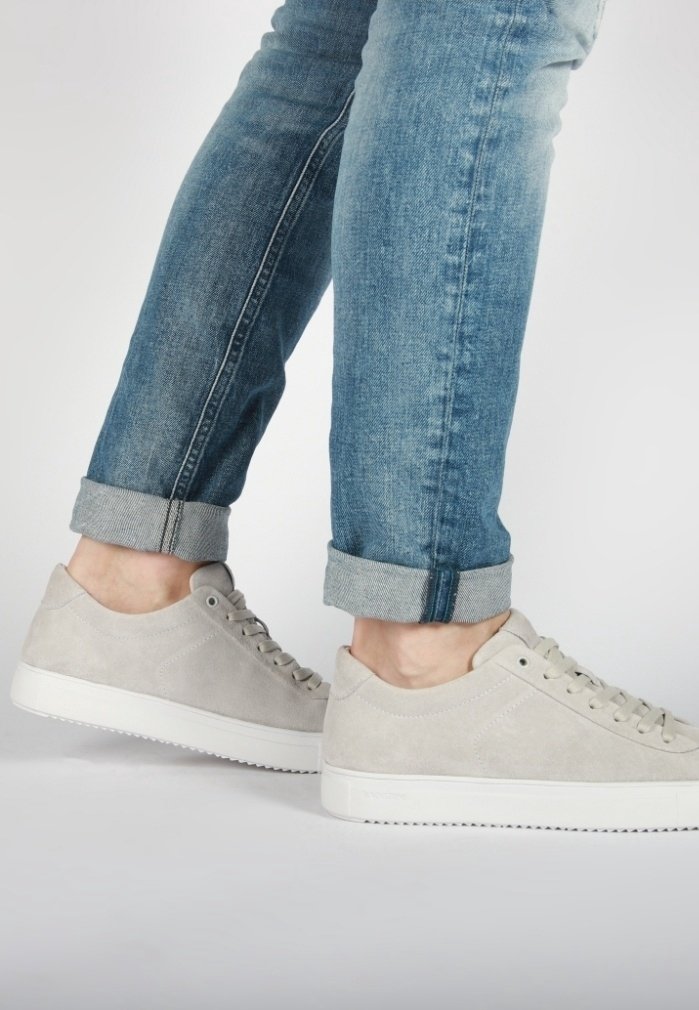 Personne portant des baskets gris clair et un jean bleu retroussé, se tenant sur la pointe des pieds devant un fond blanc uni.
