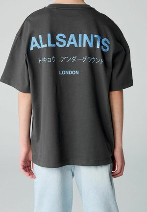 Person iført sort AllSaints t-shirt med blå tekst på engelsk og japansk, kombineret med lyseblå jeans, vist bagfra.