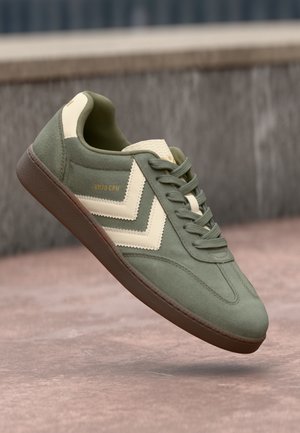 Green suede sneaker with cream accents, featuring a textured side design and round laces. Tan rubber sole and branding on the side.