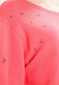 Maglione a maglia corallo con forme geometriche ricamate in nero sulle spalle. Il tessuto ha una consistenza morbida e un design con una vestibilità rilassata.