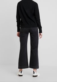 Persona que lleva pantalones vaqueros anchos negros, camiseta de manga larga negra y zapatillas blancas, de pie de espaldas contra un fondo liso.