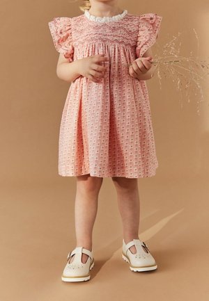 Roze katoenen jurk met bloemenpatroon, gerimpelde lijfje en gerafelde mouwen. Kind draagt beige schoenen met uitsparingen en een gesp.