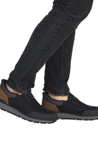 Zapatos negros de tipo slip-on con acentos marrones, parte superior sintética texturizada, suela de goma y paneles elásticos laterales, combinados con jeans oscuros.
