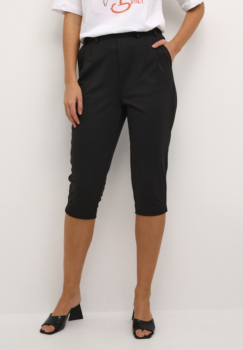 Pantalons noirs arrivant aux genoux avec une coupe ajustée, dotés de poches latérales et d'une texture de tissu lisse. Associés à un t-shirt graphique blanc et des talons noirs.