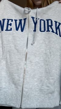Haut de survêtement zippé gris avec l'inscription bleue "NEW YORK", deux poches avant, cordons de serrage sur la capuche et poignets côtelés en bas.