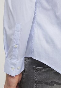 Camicia di cotone azzurro chiaro con una texture liscia, dotata di polsini con bottoni e un taglio su misura. Vista da dietro, abbinata a jeans grigi.