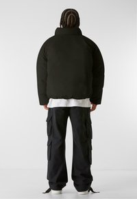 Giacca a vento nera con colletto alto, abbinata a pantaloni cargo neri larghi con molte tasche e una camicia bianca a strati sotto.