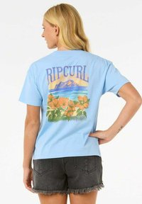 Lyseblå bomulds T-shirt med "RIPCURL" tekst og grafik af blomster, bjerge og hav på bagsiden, parret med mørke denim shorts.