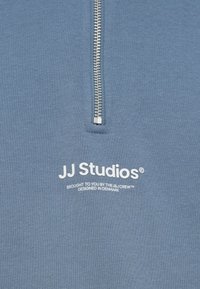 Μπλε φούτερ με φερμουάρ μέχρι τη μέση. Διαθέτει μαλακή υφή, λευκό εκτυπωμένο λογότυπο "JJ Studios" και επιπλέον κείμενο κοντά στο φερμουάρ.