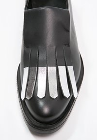 Chaussure en cuir noir avec une finition matte distincte, ornée de franges argentées à l'avant, créant un design et une texture uniques.