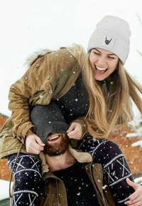 Gray knitted beanie with a logo patch, tan parka with fur trim, black sweater, patterned leggings, and snow in the background.