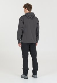Lichte grijze jas met een capuchon, lange mouwen en een zachte textuur. Draagt een zwarte broek en wandelschoenen, gezien van achteren.