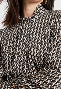 Blusa nera con motivo a fantasia che presenta un design geometrico astratto in bianco e beige, con colletto alto e tessuto testurizzato.