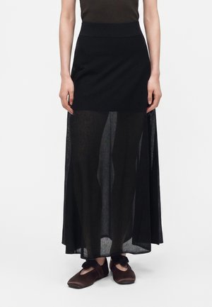 Femme portant une jupe noire taille haute avec une partie inférieure transparente et des chaussures plates marron avec des détails en nœud, debout sur un fond blanc.