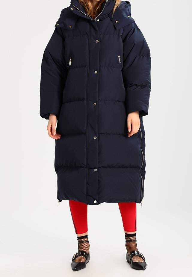 Dlouhá námořnická prošívaná bunda s kapucí, patentkami a zipovými kapsami. Texturovaná tkanina s prošívaným vzorem. Nosená přes červené legíny a černé boty.