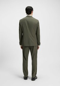 Olivgrüner Anzug mit taillierter Silhouette. Verfügt über strukturierte Schultern, eine Zwei-Knopf-Verschluss und flach geschnittene Hosen. Schwarze Schuhe.