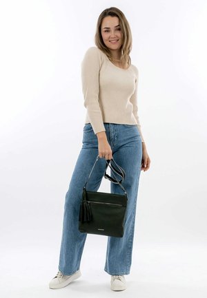 Pull beige à côtes, jeans bleus taille haute, et un sac à main en cuir noir avec un pompon, tenu sur le côté droit. Baskets blanches aux pieds.