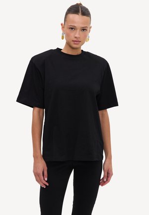Femme aux cheveux plaqués en arrière portant un t-shirt noir ample et un pantalon noir, debout devant un fond blanc uni.