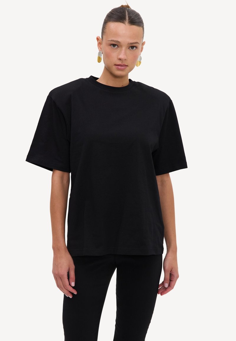 Femme aux cheveux plaqués en arrière portant un t-shirt noir ample et un pantalon noir, debout devant un fond blanc uni.