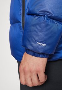 Niebieska kurtka puchowa ma rękawy o pikowanej fakturze, elastyczne mankiety oraz szczegół z białym logo "700". Widoczny zamek z przodu.