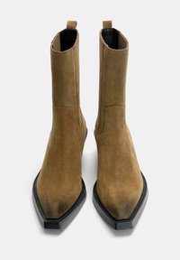 Une paire de bottes en suede tan mi-mollet avec des pointes effilées, des semelles noires, des panneaux latéraux élastiques et des tirettes en haut sur un fond blanc.