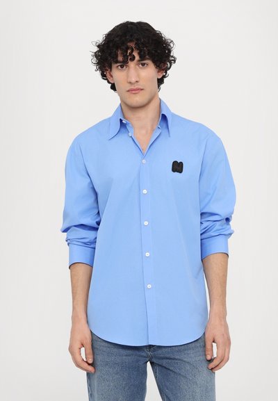 Hellblaues, knopfbares Shirt aus Baumwolle mit einem schwarzen gestickten Logo auf der linken Brust und langen Ärmeln, die an den Manschetten aufgerollt sind.