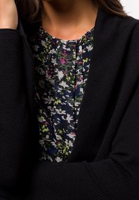 Cardigan noir texturé superposé à un chemisier floral multicolore présentant de petites fleurs blanches, roses et vertes sur un fond sombre.