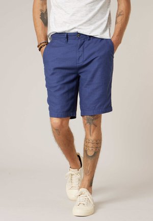 Homme portant un short bleu jusqu'aux genoux, des baskets blanches et un t-shirt blanc, avec des tatouages visibles sur les bras et les jambes.