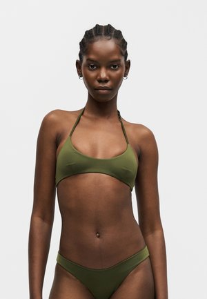 Νεαρή γυναίκα με κοτσίδες που φοράει πράσινο μπικίνι δύο τεμαχίων, κοιτάζοντας μπροστά σε απλό φωτεινό φόντο.