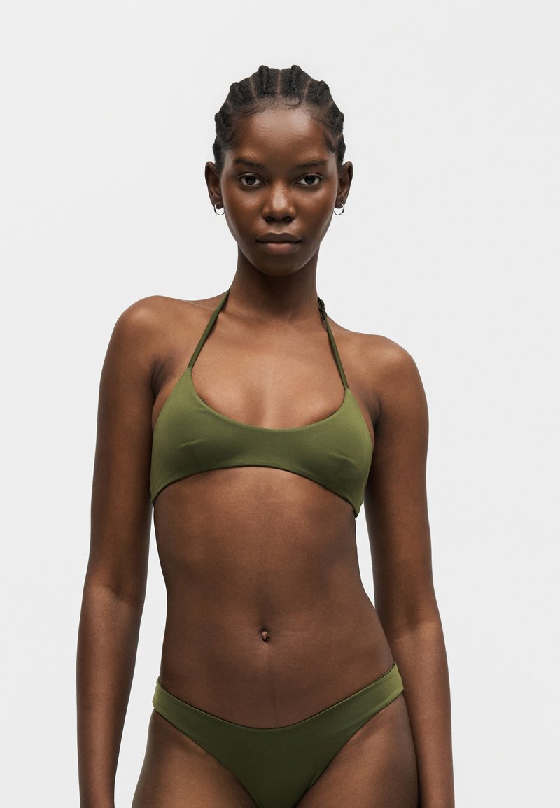 Νεαρή γυναίκα με κοτσίδες που φοράει πράσινο μπικίνι δύο τεμαχίων, κοιτάζοντας μπροστά σε απλό φωτεινό φόντο.