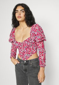 Bodysuit vermelho floral com mangas bufantes, decote em forma de barco e bordas com babados. Combinado com jeans cinza de cintura alta. O padrão apresenta pequenas flores azuis e brancas.