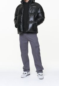 Czarna pikowana kurtka z błyszczącym wykończeniem, kapturem i przednim zamkiem błyskawicznym, noszona na czarnej bluzie z kapturem. Połączona z szarymi spodniami typu cargo i białymi trampkami.