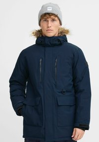 Tamsiai mėlynas šiltas parka su kailiniu gobtuvu, dviem užtrauktukais uždarytomis krūtinės kišenėmis ir didelėmis priekinėmis kišenėmis. Dėvima su pilku kepurėliu.