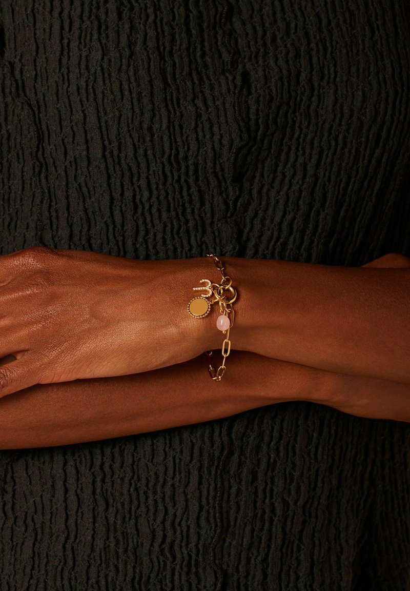 Gouden kettingarmband met ronde hanger, roze steen en gekoppelde accenten; gedragen op een gestructureerd donkere ondergrond.