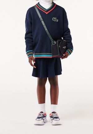Niña vistiendo un suéter azul marino de Lacoste con ribetes multicolores, falda a juego, cuello alto blanco, zapatillas deportivas y bolso cruzado de Lacoste.