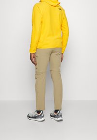 En person klädd i en gul fleecjacka och beige cargobyxor, vänd mot en vägg, grå och vita sneakers med svarta strumpor synliga.