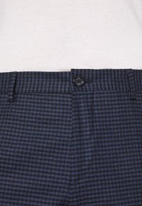 I pantaloni a scacchi blu navy presentano una chiusura con bottone nero, un tessuto strutturato e un design su misura con pieghe nette e tasche.
